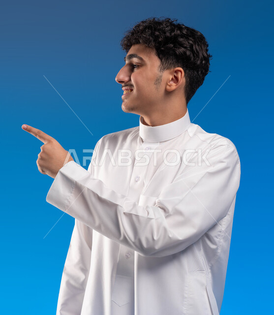 النظر إلى شيء ما بتعابير الفرح والسرور، التأمل و التخطيط للمستقبل، بورتريه مقرب لشاب عربي خليجي سعودي مبتسم يرتدي الثوب التقليدي يشير بإصبعه إلى شيء محدد، حركات وإيماءات مختلفة، خلفية زرقاء .