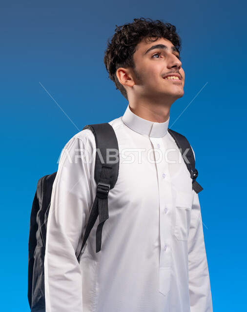 رؤية مستقبلية للتعليم، إيماءات التفكير والتأمل، الاستعداد والذهاب إلى المدرسة، تطوير المناهج التعليمية في السعودية, بورتريه لشاب عربي سعودي خليجي يرتدي الثوب المدرسي يحمل الحقيبة والكتب المدرسية ينظر إلى شيء ما, خلفية زرقاء .