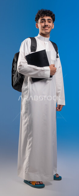 تطوير المناهج التعليمية في السعودية, النظر إلى الكاميرا بإيماءات الاستعداد، متابعة الدروس والواجبات، بورتريه لشاب عربي سعودي خليجي مبتسم يرتدي الثوب المدرسي يحمل الحقيبة والكتب المدرسية, أهمية التعليم في بناء المستقبل, صورة الجسم بالطول الكامل، خلفية زرقاء .