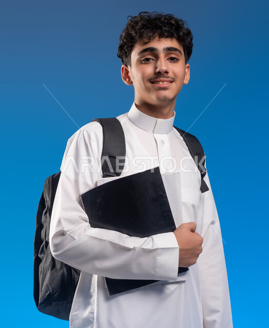 Looking at the camera with gestures of readiness, following lessons and assignments, developing educational curricula in Saudi Arabia, portrait of a smiling Saudi Gulf Arab young man wearing a school uniform carrying a bag and textbooks, the beginning of the new school year, the importance of education in building the future, blue background