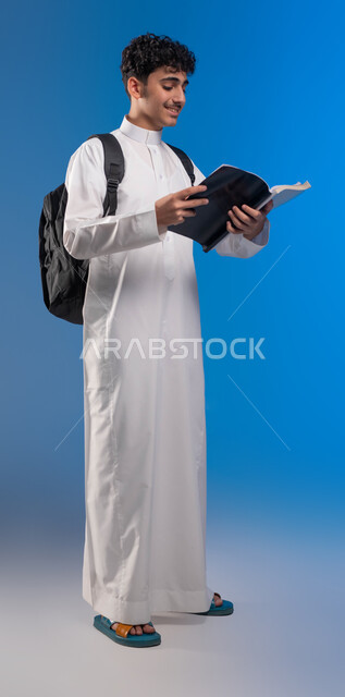 Gestures of diligence and preparing lessons, following up on lessons and assignments, developing educational curricula in Saudi Arabia, portrait of a Saudi Gulf Arab young man wearing a school dress carrying a bag and textbooks, the beginning of the new school year, full-length body portrait, blue background