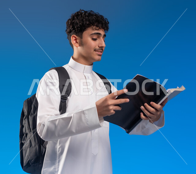 Following up on lessons and assignments, developing educational curricula in Saudi Arabia, a portrait of a Saudi Gulf Arab young man wearing a school dress carrying a bag and textbooks, the beginning of the new school year, gestures of diligence and preparing lessons, blue background
