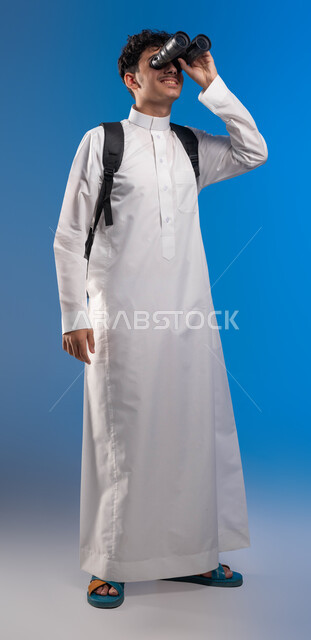 رؤية الاشياء البعيدة عبر المنظار، حب المغامرة والاستكشاف، بورتريه لطالب عربي خليجي سعودي مبتسم يرتدي الثوب التقليدي وحقيبة الظهر ينظر من خلال منظار الرؤية, التخطيط المستقبلي للأهداف والطموحات، صورة الجسم بالطول الكامل، خلفية زرقاء
