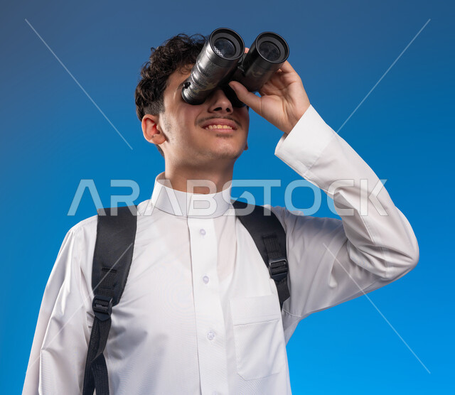 رؤية الاشياء البعيدة عبر المنظار، حب المغامرة والاستكشاف، بورتريه مقرب لفتى عربي خليجي سعودي مبتسم يرتدي الثوب التقليدي وحقيبة الظهر ينظر من خلال منظار الرؤية, التخطيط المستقبلي للأهداف والطموحات، خلفية زرقاء