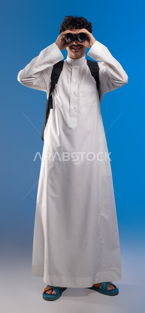 التخطيط المستقبلي للأهداف والطموحات، بورتريه لطالب عربي خليجي سعودي مبتسم يرتدي الثوب التقليدي وحقيبة الظهر ينظر من خلال منظار الرؤية, رؤية الاشياء البعيدة عبر المنظار، حب المغامرة والاستكشاف، صورة الجسم بالطول الكامل، خلفية زرقاء