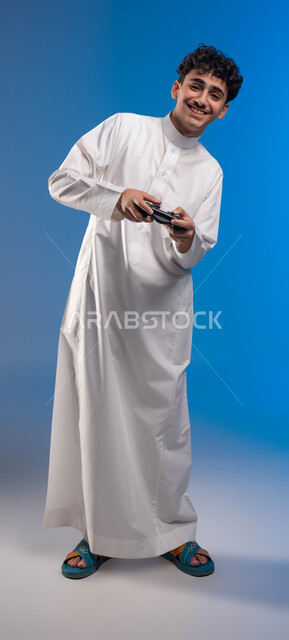 أجهزة تكنولوجية حديثة، دمج العالم الالكتروني مع التسلية، بورتريه لفتى عربي خليجي سعودي مبتسم يرتدي الثوب التقليدي يمسك بيده عصا التحكم يقوم باللعب في ألعاب الفيديو الترفيهية، تعابير تدل على الحماس والإنسجام، صورة الجسم بالكامل، خلفية زرقاء