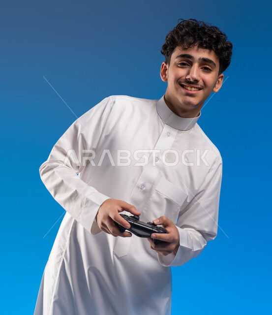 دمج المتعة والتسلية مع العالم الالكتروني، بورتريه مقرب لفتى عربي خليجي سعودي مبتسم يرتدي الثوب التقليدي يمسك بيده عصا التحكم يقوم باللعب في ألعاب الفيديو الترفيهيه، تعابير تدل على الحماس والإنسجام، أجهزة تكنولوجية وتقنيات حديثة، خلفية زرقاء