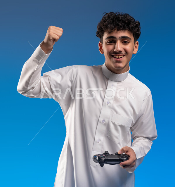 ايماءات يد تدل على الانتصار والفوز، دمج العالم الالكتروني مع التسلية والمتعة، بورتريه مقرب لفتى عربي خليجي سعودي مبتسم يرتدي الثوب التقليدي يمسك بيده عصا التحكم يقوم باللعب في ألعاب الفيديو الترفيهية، أجهزة تكنولوجية وتقنيات حديثة، خلفية زرقاء