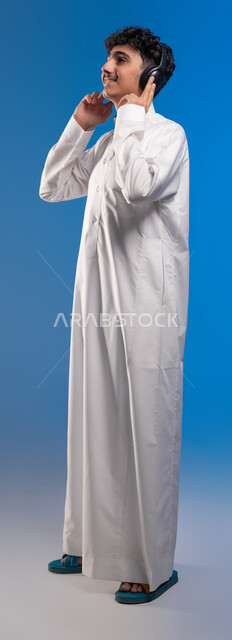 Spending enjoyable times listening to singing talents, the concept of listening and enjoying using a headphone, a portrait of a Saudi Arabian Gulf boy wearing a traditional dress and wearing a headset, facial gestures indicating integration and joy, full-length body image, blue background