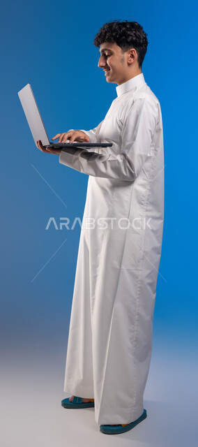 الضغط على أزرار لوحة المفاتيح، تصفح الانترنت ووسائل التواصل الاجتماعي، بورتريه جانبي لفتى عربي خليجي سعودي مبتسم يرتدي الثوب التقليدي يقوم باستخدام الحاسوب المحمول، احتراف وظائف الحاسوب وتكنولوجيا المعلومات، صورة الجسم بالكامل، خلفية زرقاء