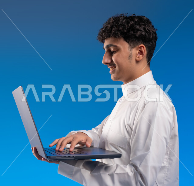 تصفح الانترنت ووسائل التواصل الاجتماعي، بورتريه جانبي مقرب لفتى عربي خليجي سعودي مبتسم يرتدي الثوب التقليدي يقوم باستخدام الحاسوب المحمول، الضغط على أزرار لوحة المفاتيح، احتراف وظائف الحاسوب وتكنولوجيا المعلومات، خلفية زرقاء