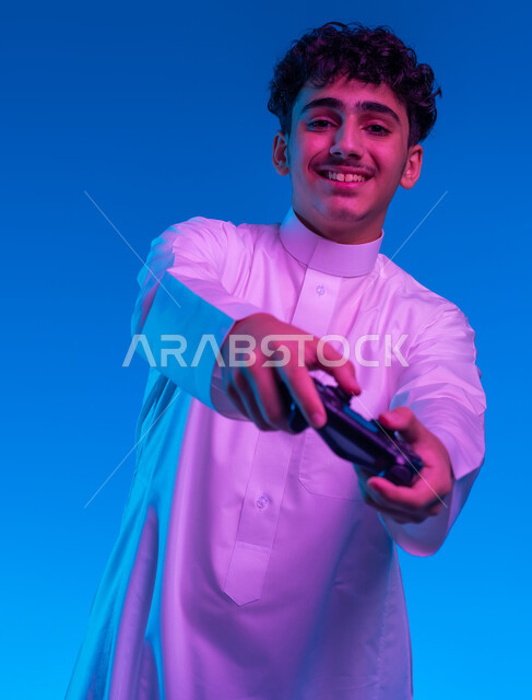 دمج العالم الالكتروني مع المتعة والتسلية، بورتريه مقرب لفتى عربي خليجي سعودي مبتسم يرتدي الثوب التقليدي يمسك بيده عصا التحكم يقوم باللعب في ألعاب الفيديو الترفيهية، تعابير تدل على الحماس والإنسجام، أجهزة تكنولوجية وتقنيات حديثة، خلفية زرقاء