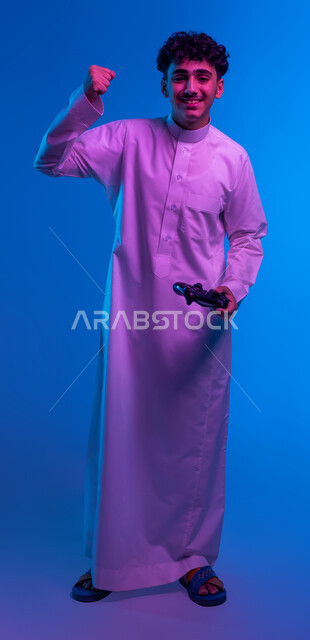 Gestures indicating triumph and victory, merging the imaginary world with entertainment, portrait of a smiling Saudi Arabian Gulf boy wearing a traditional dress holding a joystick in his hand playing entertainment video games, modern technological devices, full body image, blue background