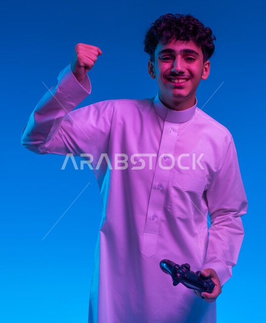 تعابير تدل على الانتصار والفوز، دمج العالم الالكتروني مع التسلية، بورتريه مقرب لفتى عربي خليجي سعودي مبتسم يرتدي الثوب التقليدي يمسك بيده عصا التحكم يقوم باللعب في ألعاب الفيديو الترفيهية، أجهزة تكنولوجية حديثة، خلفية زرقاء