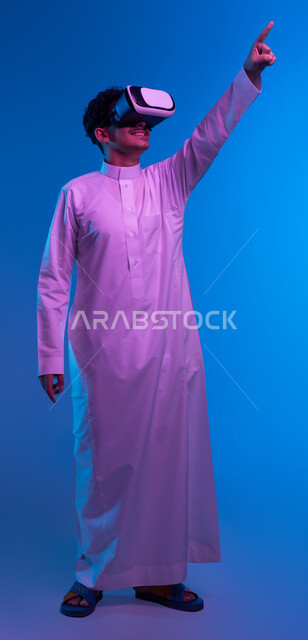 محاكاة العالم الخيالي، رؤية مستقبلية بعيدة، بورتريه لفتى عربي خليجي سعودي مبتسم يرتدي الثوب التقليدي ونظارات الواقع الافترضي ثلاثية الابعاد يشير بإصبع السبابة الى مكان بعيد، أجهزة تكنولوجية وتقنيات حديثة، صورة الجسم بالطول الكامل، خلفية زرقاء