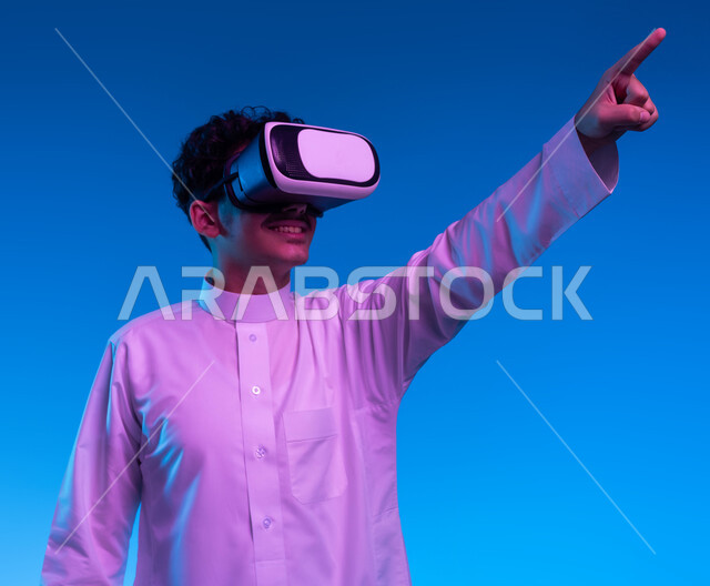 رؤية مستقبلية بعيدة، بورتريه مقرب لفتى عربي خليجي سعودي مبتسم يرتدي الثوب التقليدي ونظارات الواقع الافترضي ثلاثية الابعاد يشير بإصبع السبابة الى مكان بعيد، أجهزة تكنولوجية وتقنيات حديثة، محاكاة العالم الخيالي، خلفية زرقاء