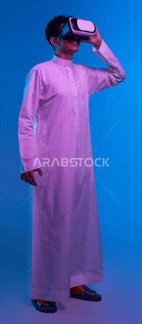 تعابير وايماءات تدل على الاندماج والمتعة, بورتريه لفتى عربي خليجي سعودي مبتسم يرتدي الثوب التقليدي يقوم بمشاهدة أفلام عبر نظارات الواقع الافتراضي ثلاثية الأبعاد, محاكاة العالم الخيالي والتفاعل مع الفيلم، صورة الجسم بالطول الكامل، خلفيه زرقاء