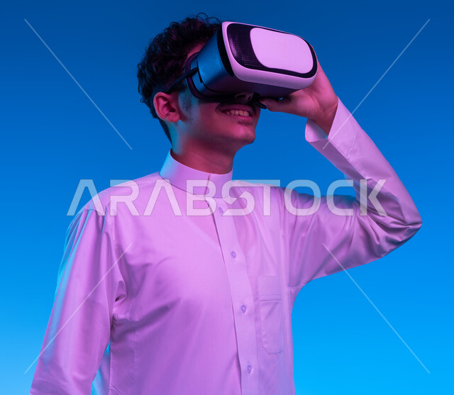 التفاعل مع الفيلم ومحاكاة العالم الخيالي، بورتريه مقرب لفتى عربي خليجي سعودي مبتسم يرتدي الثوب التقليدي يقوم بمشاهدة أفلام عبر نظارات الواقع الافتراضي ثلاثية الأبعاد, ايماءات تدل على الاندماج والمتعة, خلفيه زرقاء