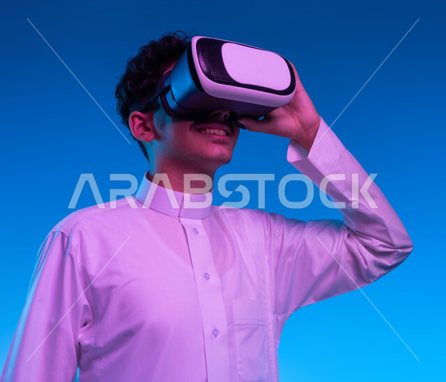 محاكاة العالم الخيالي والتفاعل مع الفيلم، بورتريه مقرب لفتى عربي خليجي سعودي مبتسم يرتدي الثوب التقليدي يقوم بمشاهدة أفلام عبر نظارات الواقع الافتراضي ثلاثية الأبعاد, ايماءات تدل على الاندماج والمتعة, خلفيه زرقاء
