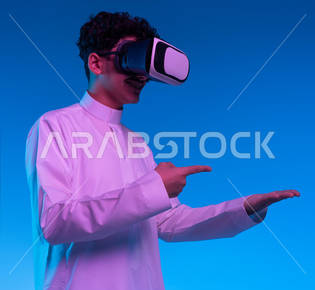 استخدام التقنيات الحديثة والمتطورة, بورتريه جانبي مقرب لفتى عربي خليجي سعودي يرتدي الثوب التقليدي ونظارات الواقع الافتراضي ثلاثية الابعاد يشير باصبع السبابة نحو يده الاخرى، محاكاة العالم الخيالي والتفاعل معه، خلفيه زرقاء