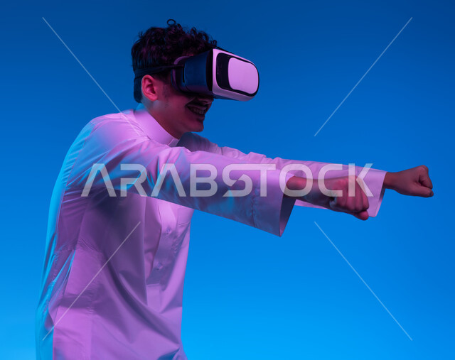 تقنيات وأجهزة حديثة، محاكاة ركوب دراجة ثري دي في العالم الخيالي، بورتريه جانبي مقرب لولد عربي خليجي سعودي يرتدي الثوب التقليدي ونظارة الواقع الإفتراضي ثلاثية الأبعاد يقوم باللعب في ألعاب الفيديو، إيماءات يدين تدل على السعادة بألعاب قيادة الدراجات، خلفية زرقاء