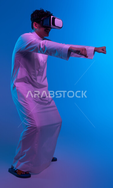 إيماءات يدين تدل على قيادة الدراجات، أجهزة وتقنيات تكنولوجية، محاكاة ركوب دراجة ثري دي في العالم الخيالي، بورتريه لولد عربي خليجي سعودي يرتدي الثوب التقليدي ونظارة الواقع الإفتراضي ثلاثية الأبعاد يقوم باللعب في ألعاب الفيديو، صورة الجسم بالكامل، خلفية زرقاء