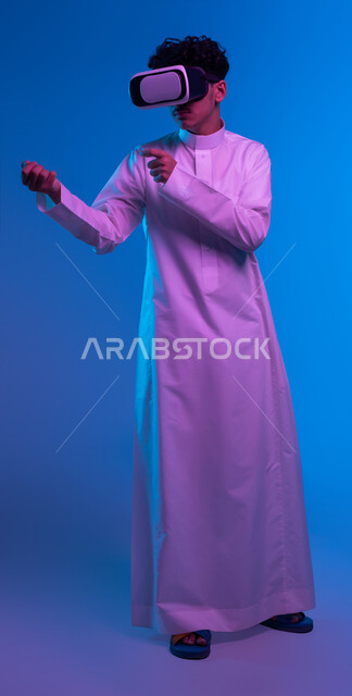 صورة الجسم بالكامل، تفريغ الطاقة السلبية، ممارسة رياضة الدفاع عن النفس بتقنيات تكنولوجية، التدرب على الحركات القتالية عبر ألعاب الفيديو، بورتريه لفتى عربي خليجي سعودي يرتدي الثوب التقليدي ونظارة الواقع الافتراضي بإيماءات الملاكمة والمصارعة، خلفية زرقاء