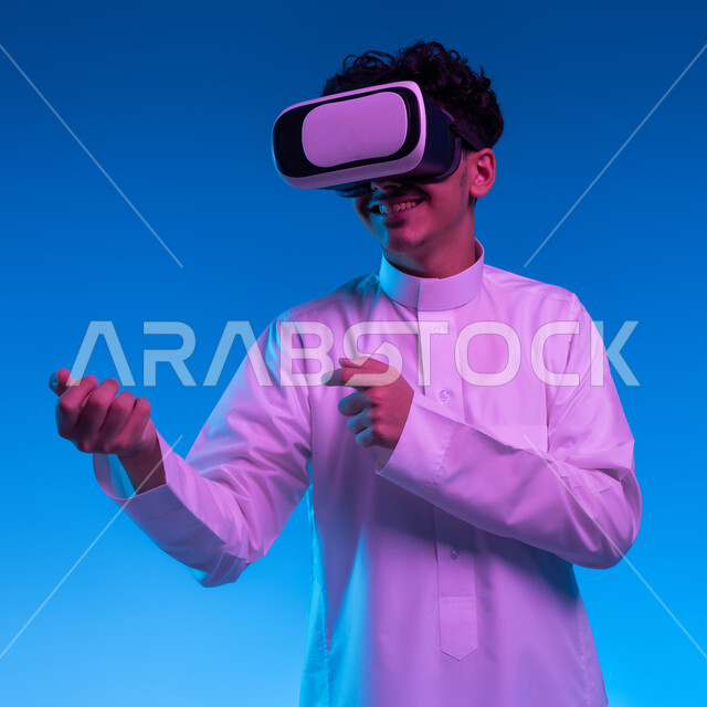 التدرب على الحركات القتالية من خلال ألعاب الفيديو، تفريغ الطاقة السلبية، ممارسة رياضة الدفاع عن النفس بتقنيات تكنولوجية، بورتريه مقرب لفتى عربي خليجي سعودي مبتسم يرتدي الثوب التقليدي ونظارات الواقع الافتراضي بإيماءات الملاكمة والمصارعة، خلفية زرقاء