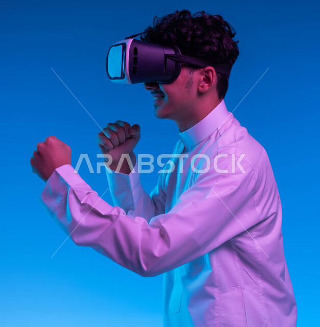 تفريغ الطاقة السلبية، ممارسة رياضة الدفاع عن النفس بتقنيات تكنولوجية، التدرب على الحركات القتالية من خلال ألعاب الفيديو، بورتريه جانبي مقرب لفتى عربي خليجي سعودي يرتدي الثوب التقليدي ونظارات الواقع الافتراضي بإيماءات الملاكمة والمصارعة، خلفية زرقاء