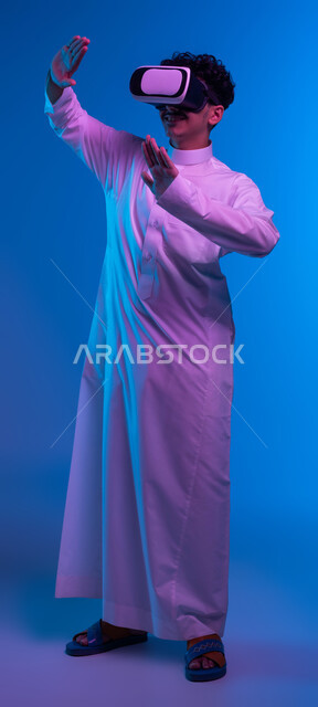 الاندماج ومحاولة امساك شيء ما, التفاعل ومحاكاة العالم الخيالي, بورتريه لفتى عربي خليجي سعودي يرتدي الثوب التقليدي و نظارات الواقع الافتراضي بايماءات الدفاع عن النفس، تقنيات حديثة وأجهزة تكنولوجية، صورة الجسم بالطول الكامل، خلفية زرقاء