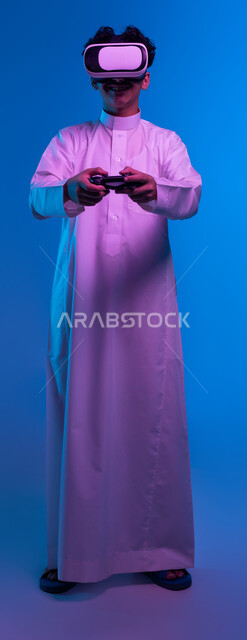 تقنيات حديثة وأجهزة تكنولوجية، بورتريه لفتى عربي خليجي سعودي يرتدي الثوب التقليدي ونظارة الواقع الإفتراضي يمسك بيده عصا التحكم يقوم باللعب في ألعاب الفيديو الترفيهية، دمج العالم الخيالي مع المتعة والتسلية، صورة الجسم بالكامل، خلفية زرقاء