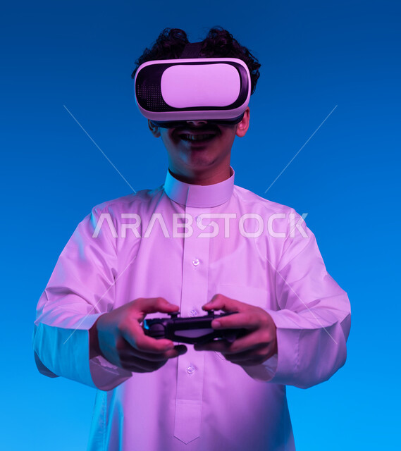 دمج العالم الخيالي مع المتعة والتسلية، بورتريه مقرب لفتى عربي خليجي سعودي مبتسم يرتدي الثوب التقليدي ونظارة الواقع الإفتراضي يمسك بيده عصا التحكم يقوم باللعب في ألعاب الفيديو الترفيهية، أجهزة تكنولوجية وتقنيات حديثة، خلفية زرقاء