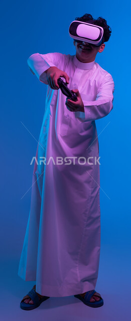 أجهزة تكنولوجية وتقنيات حديثة، بورتريه لفتى عربي خليجي سعودي يرتدي الثوب التقليدي ونظارة الواقع الإفتراضي يمسك بيده عصا التحكم يقوم باللعب في ألعاب الفيديو الترفيهية، دمج العالم الخيالي مع المتعة والتسلية، صورة الجسم بالكامل، خلفية زرقاء