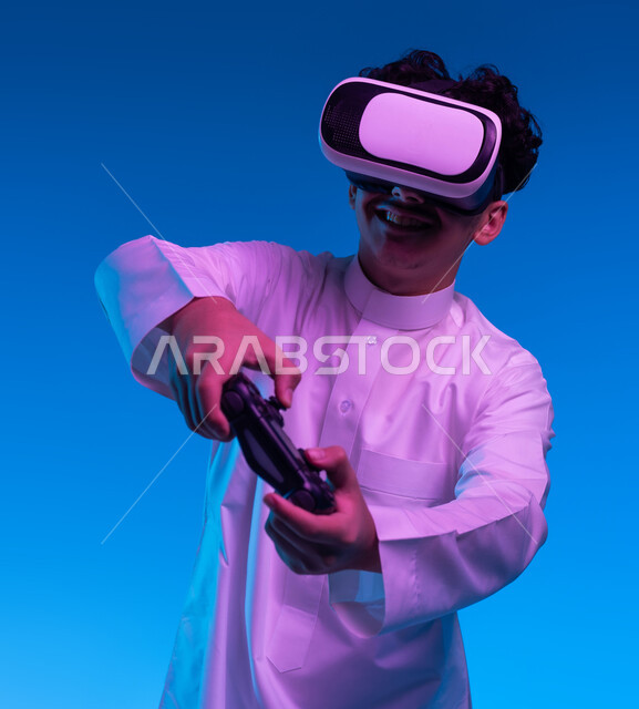 دمج المتعة والتسلية مع العالم الخيالي، بورتريه مقرب لفتى عربي خليجي سعودي مبتسم يرتدي الثوب التقليدي ونظارة الواقع الإفتراضي يمسك بيده عصا التحكم يقوم باللعب في ألعاب الفيديو الترفيهية، أجهزة تكنولوجية وتقنيات حديثة، خلفية زرقاء