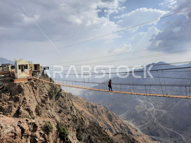 فعالية السير على الجسر الخشبي المعلق في حديقة الأمير حسام، القمم والمرتفعات الجبلية الصخرية في الباحة، سائح يمارس النشاطات الحركية المسلية وسط المناظر الطبيعة في منتزهات وحدائق المملكة، أنشطة ترفيهية ممتعة في الأماكن السياحية السعودية
