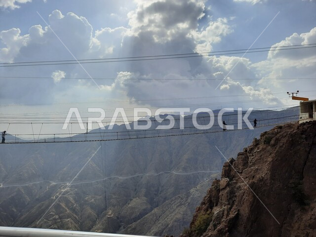 مجموعة من الأشخاص يمارسون النشاطات الحركية المسلية وسط المناظر الطبيعة في منتزهات وحدائق المملكة، فعالية السير على الجسر الخشبي المعلق في حديقة الأمير حسام، القمم والمرتفعات الجبلية الصخرية في الباحة، أنشطة ترفيهية ممتعة في الأماكن السياحية السعودية