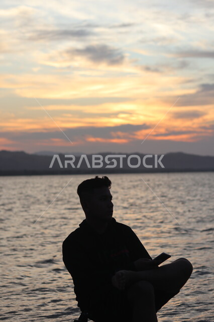 الشعور بالاكتئاب والوحدة، الحاجة إلى التأمل والاسترخاء أمام البحر وقت غروب الشمس، الاستمتاع بأوقات الفراغ في المناطق الساحليه السياحيه, شاب عربي خليجي سعودي يجلس على شاطئ البحر ويحمل بيده الهاتف المحمول وينظر الى شيء ما، سيلوليت