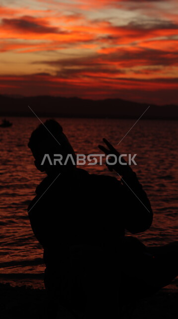 الاسترخاء والتأمل أمام البحر وقت غروب الشمس، صورة ظليه لشاب عربي خليجي سعودي يجلس على شاطئ البحر بإيماءات النصر والفوز، الاستمتاع بأوقات الفراغ في المناطق الساحليه السياحيه, سيلوليت