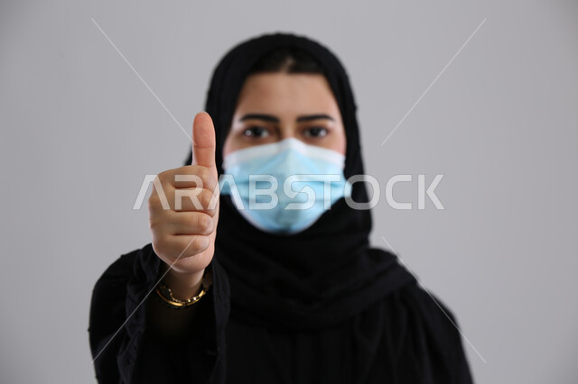 رفع الإبهام نحو الأعلى للحرص على الوقايه والسلامه, ايماءات اتباع الاحترازات الوقائيه لمكافحة فيروس كورونا ، بورتريه مقرب لإمرأة عربية سعودية خليجية محجبة ترتدي الكمامة لتجنب انتشار العدوى ، خلفية رمادية