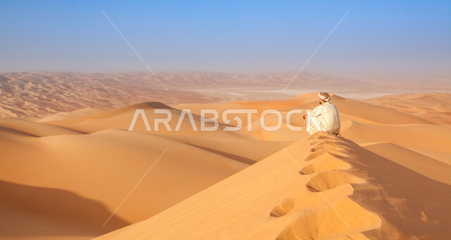 الاستمتاع بالمناظر الهادئة الطبيعيه في وقت النهار, رجل عربي خليجي يرتدي الزي التقليدي يجلس فوق الكثبان الرملية في الصحراء العربية, المناطق الصحراوية في شبه الجزيرة, التضاريس والرمال الذهبية الناعمة بالأماكن السياحية في الخليج العربي.