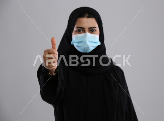 Maintaining safety and avoiding the spread of infection from Covid-19, feeling accepted and satisfied, taking preventive precautions against the Corona virus, close-up portrait of a veiled Saudi Arabian Gulf woman wearing a mask, raising her thumb up with a quality sign, gray background