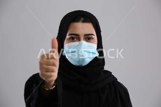 إجراء الاحترازات الوقائية من فايروس كورونا ،الشعور بالقبول والرضا , بورتريه مقرب لإمرأة عربية سعودية خليجية محجبة ترتدي الكمامة ترفع اصبع الإبهام للأعلى بعلامة الجودة , الحفاظ على السلامة و تجنب انتشار العدوى من كوفيد 19 ، خلفية رمادية