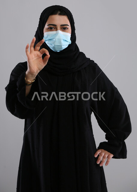 Precaution and prevention of the Corona virus, feeling accepted and satisfied, portrait of a veiled Saudi Arabian Gulf woman wearing a mask, raising her hand upwards with a quality sign, maintaining safety and avoiding the spread of infection from Covid 19, gray background