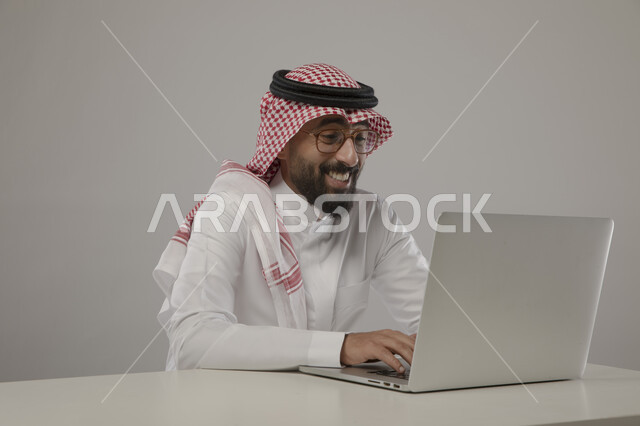 الجلوس امام الحاسوب المحمول وتصفح مواقع التواصل بايماءات السرور والسعادة، انجاز مهام العمل عن بعد، بورتريه لرجل اعمال عربي خليجي سعودي يرتدي الزي التقليدي والنظارة الطبية ، مفهوم ادارة الاعمال وتحقيق اهداف الشركة، مهن ووظائف مكتبية سعودية