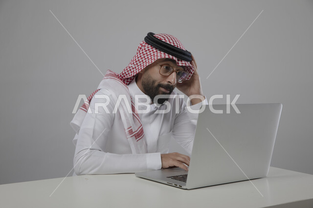 الجلوس امام الحاسوب المحمول بايماءات الحيرة والتفكير، انجاز مهام العمل عن بعد، بورتريه لرجل اعمال عربي خليجي سعودي يرتدي الزي التقليدي والنظارة الطبية وينظر الى اللابتوب بتركيز، مفهوم ادارة الاعمال وتحقيق اهداف الشركة، مهن ووظائف مكتبية سعودية