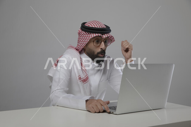 انجاز مهام العمل عن بعد، بورتريه لرجل اعمال عربي خليجي سعودي يرتدي الزي التقليدي والنظارة الطبية ويجلس امام الحاسوب المحمول بايماءات الدهشة والصدمة والذهول، مفهوم ادارة الاعمال وتحقيق اهداف الشركة، مهن ووظائف مكتبية سعودية