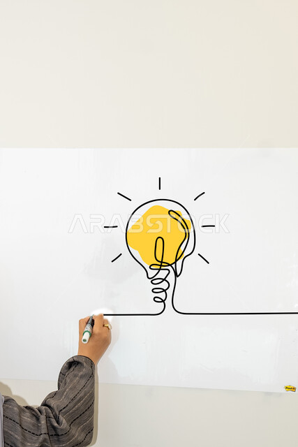 مفهوم الذكاء والعبقرية، أفكار تطويرية مميزة، التفكير مع الالهام والابتكار، تحقيق الانجازات والمهامات، صورة مقربة ليد شابة عربية خليجيه سعودية تقوم برسم لوحة فنية للمصباح الكهربائي الضوئي, خلفية بيضاء