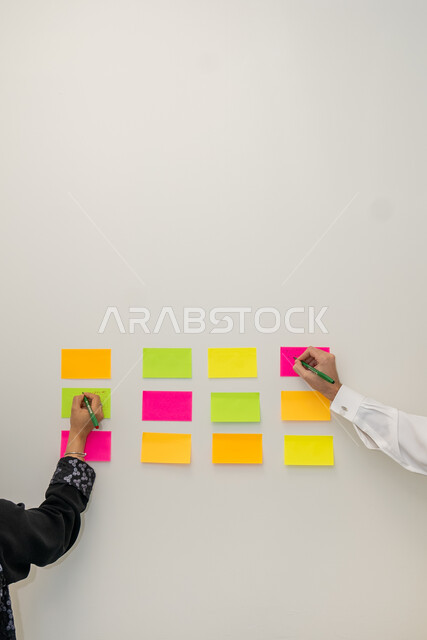 كتابة الاهداف المستقبيله و الافكار الجديده والذكيه، امرأة عربية خليجيه سعودية و شاب سعودي يقومان بتدوين الملاحظات والمهام ، توزيع الاعمال المطلوبه ، نوتات ملونه معلقه على الجدران