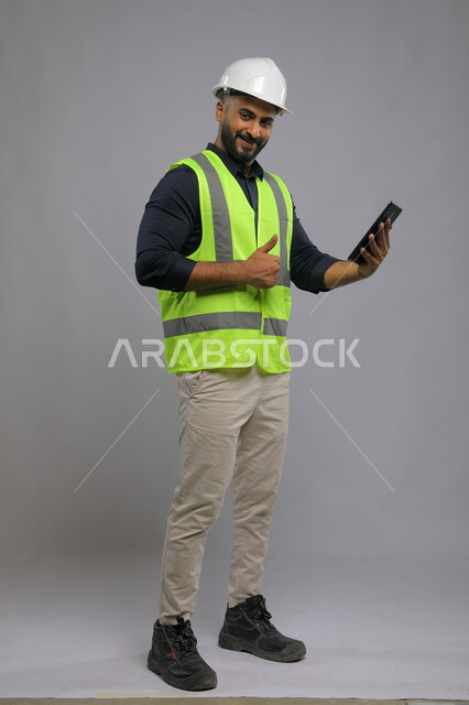 مجال الانشاءات والمشاريع الهندسية, إيماءات النظر إلى الكاميرا ورفع إصبع الإبهام نحو الأعلى بعلامة الجودة, بورتريه لمهندس مدني عربي خليجي سعودي يبتسم يرتدي الخوذة وسترة الحماية يستخدم الهاتف المحمول، صورة الجسم بالطول الكامل,  خلفية رمادية.