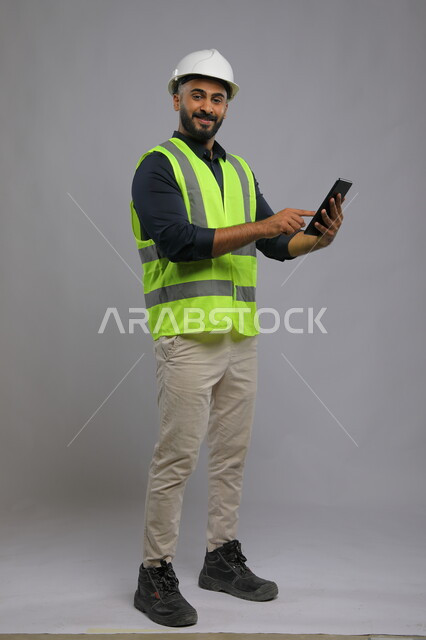 البحث عن المخططات الهندسيه عبر الجوال, دمج العمل مع التقنية و التكنولوجيا، إيماءات السرور والنظر إلى الكاميرا, بورتريه لمهندس معماري عربي خليجي سعودي يبتسم يرتدي الخوذة وسترة الحماية يستخدم الهاتف المحمول، صورة الجسم بالطول الكامل,  خلفية رمادية.